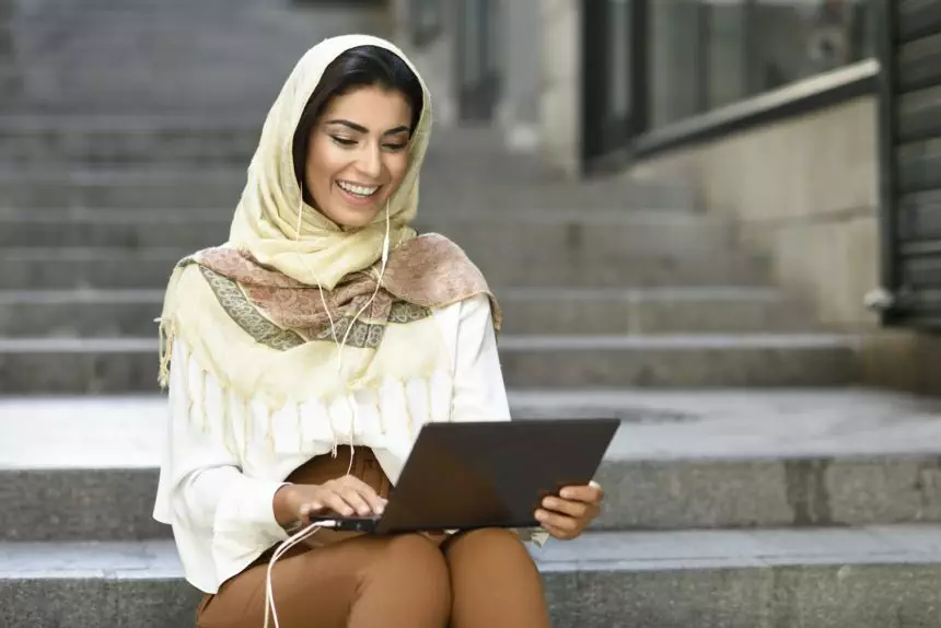 Spain, Granada, young muslim woman wearing hijab using laptop sitting on urban steps